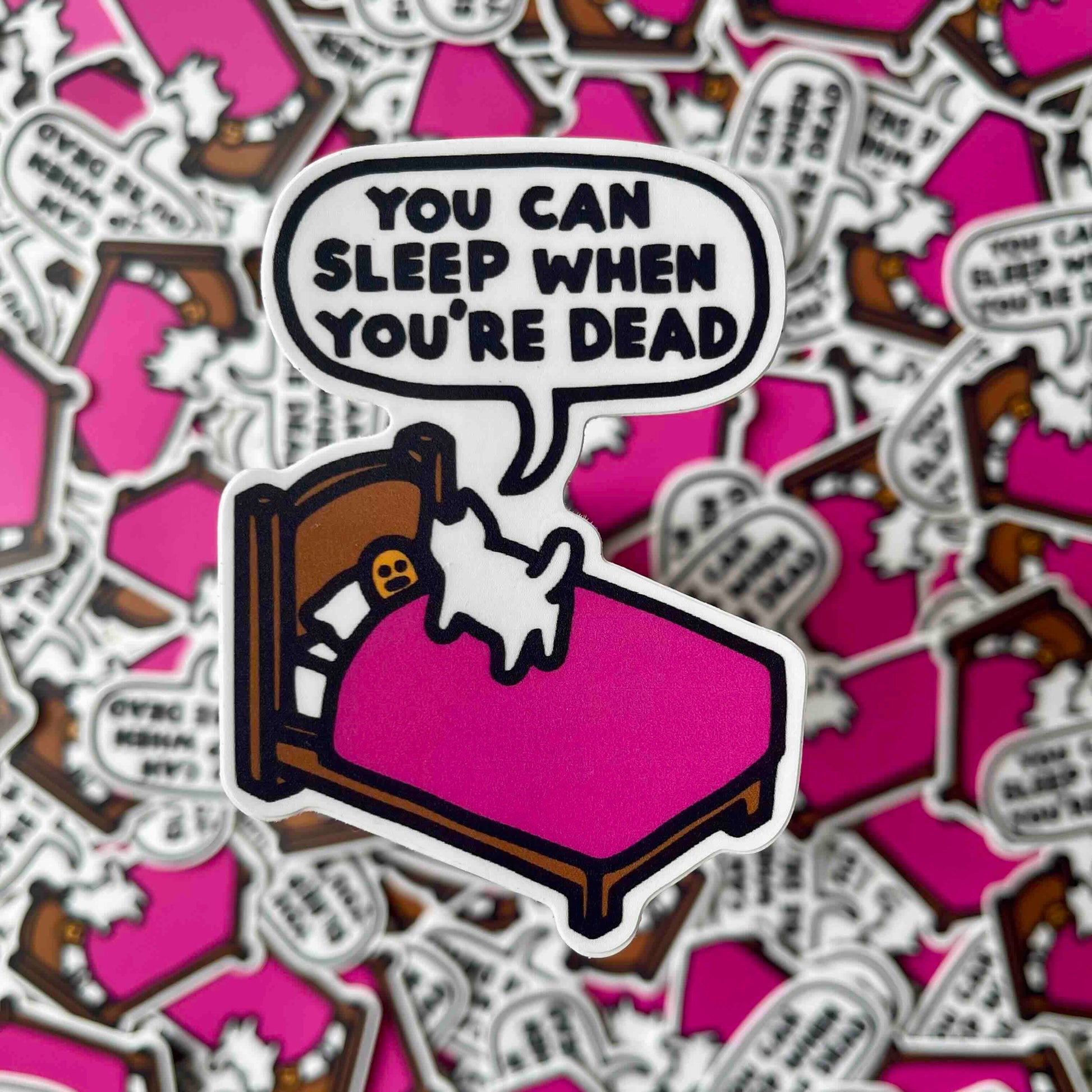 A die-cut sticker featuring a cartoon person lying in a bed with cat standing on them with a bright pink blanket and a speech bubble that reads 'YOU CAN SLEEP WHEN YOU'RE DEAD'. The sticker is placed on top of a pile of identical stickers, creating a busy background.