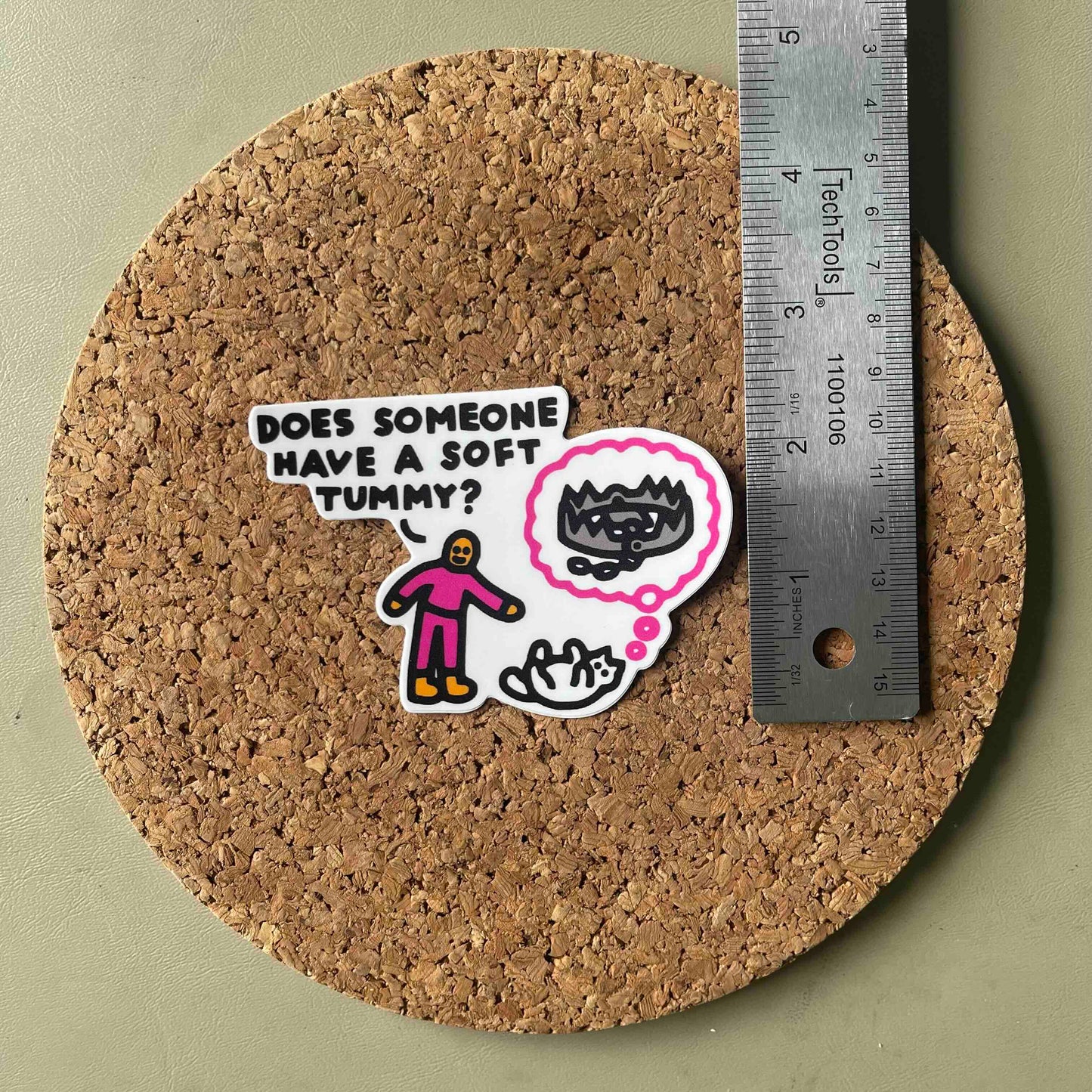 A die-cut sticker with a cartoon figure and text reading 'DOES SOMEONE HAVE A SOFT TUMMY?' shown on a round cork coaster next to a metal ruler for scale, on a muted green background.